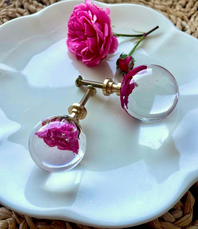 May include: Two spherical cabinet knobs featuring clear resin encasing vibrant pink rose petals. Each knob has a metallic gold-toned base and a threaded screw attachment. A single fresh pink rose and a small bud are positioned near the knobs.
