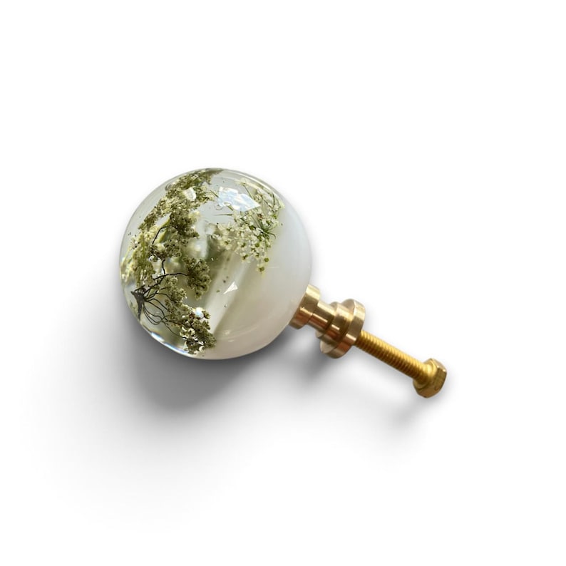 May include: A spherical cabinet knob featuring a clear resin dome with embedded dried white flowers and greenery. The knob is attached to a metallic gold-toned threaded bolt and base, providing a functional hardware piece for furniture.