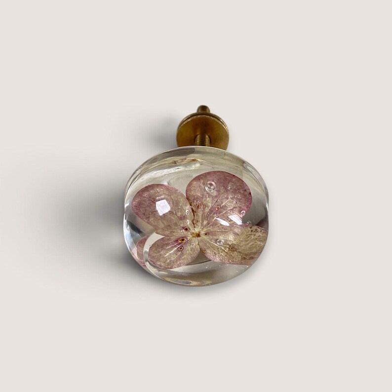 Puede incluir: Un pomo esf&eacute;rico para muebles hecho de resina transparente que contiene una flor de hortensia rosa prensada. El pomo est&aacute; unido a una base met&aacute;lica de tono lat&oacute;n y cuenta con un perno roscado corto para su montaje en muebles.