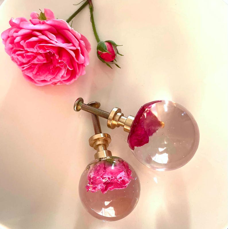 May include: Two spherical cabinet knobs featuring clear resin spheres with embedded pink rose petals. Each knob has a metallic gold-toned base and a threaded screw attachment. A single large pink rose and a small rosebud are positioned nearby.