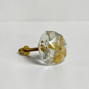 May include: A faceted, clear resin cabinet knob featuring an embedded dried flower petal in a light tan hue. The knob is attached to a metallic gold-toned threaded bolt with a matching circular washer and nut, providing a secure mounting mechanism.