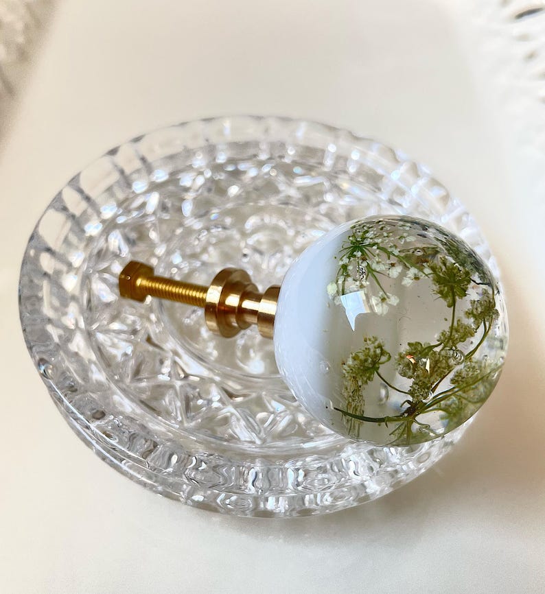 May include: A spherical cabinet knob featuring a clear resin dome with embedded white pressed flowers, attached to a gold-toned metal base and threaded bolt. The knob is positioned on a circular, clear glass decorative dish with a textured pattern.