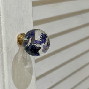 May include: A spherical clear resin cabinet knob featuring embedded small blue flowers. The knob is attached to a circular gold-toned metal base, which is mounted onto a white louvered cabinet door surface.