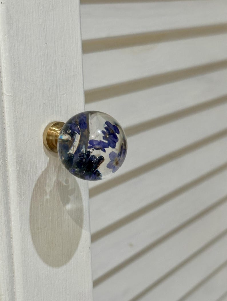 May include: A spherical clear resin cabinet knob featuring embedded small blue flowers. The knob is attached to a circular gold-toned metal base, which is mounted onto a white louvered cabinet door surface.