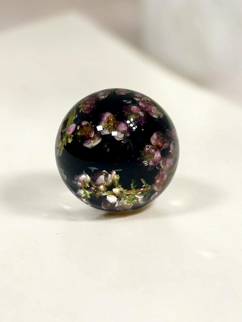 Pode incluir: Um peso de papel esf&eacute;rico de resina com uma base preta transl&uacute;cida escura incrustada com pequenas e delicadas flores roxas e brancas, juntamente com caules e folhas verdes, criando um arranjo floral suspenso dentro da esfera polida.