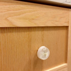 K&ouml;nnte beinhalten: Ein runder, wei&szlig;er, halbtransparenter M&ouml;belknopf mit einem eingebetteten, getrockneten, hellen Bl&uuml;tenblatt in der Mitte. Der Knopf ist an einer hellen, naturbelassenen Holzschubladenfront mit sichtbarer Maserung und flachem Paneeldesign befestigt.