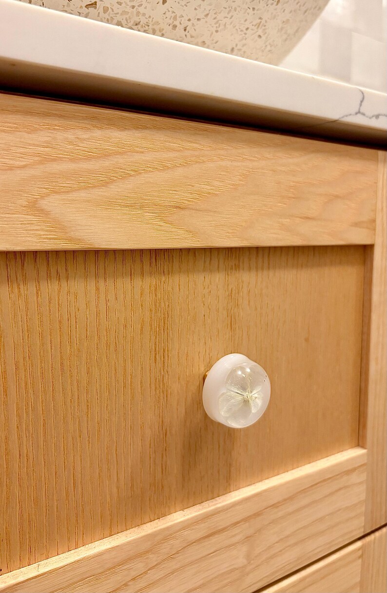 K&ouml;nnte beinhalten: Ein runder, wei&szlig;er, halbtransparenter M&ouml;belknopf mit einem eingebetteten, getrockneten, hellen Bl&uuml;tenblatt in der Mitte. Der Knopf ist an einer hellen, naturbelassenen Holzschubladenfront mit sichtbarer Maserung und flachem Paneeldesign befestigt.
