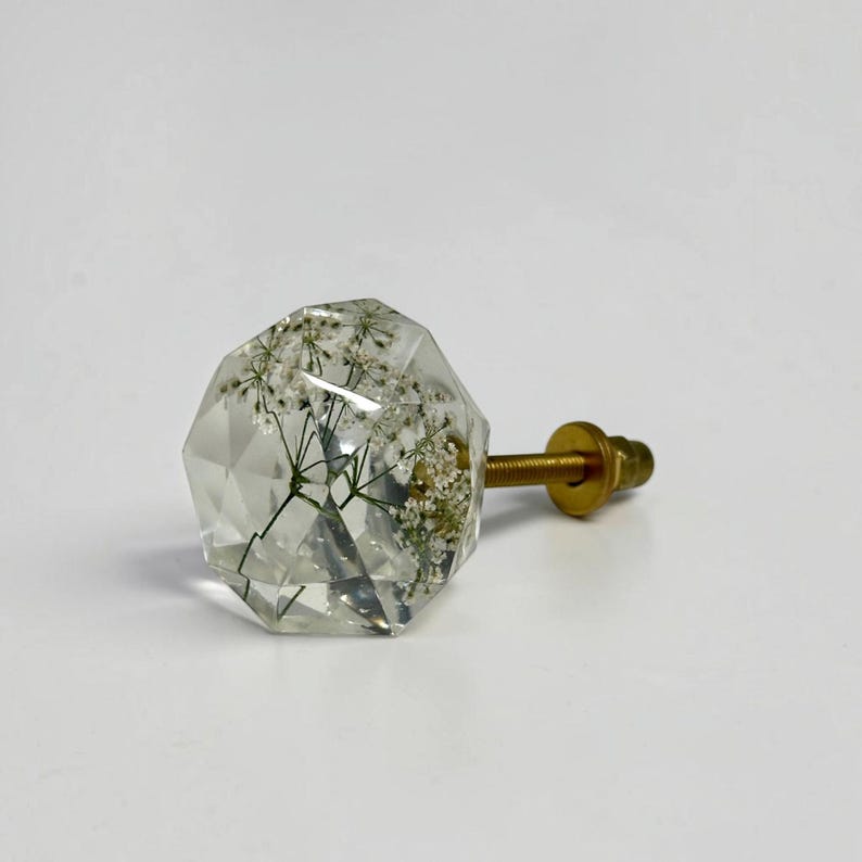 May include: A clear, faceted, geometric cabinet knob featuring embedded white dried flowers inside the resin structure. The knob is attached to a gold-toned metal screw and nut assembly, providing a functional hardware piece for furniture.