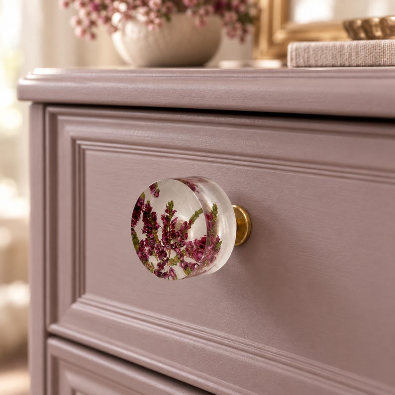 Puede incluir: Un tirador de caj&oacute;n redondo de resina transparente con flores de brezo moradas incrustadas. El tirador est&aacute; fijado a un frente de caj&oacute;n de madera de color malva con una base de metal dorado, creando un detalle decorativo.