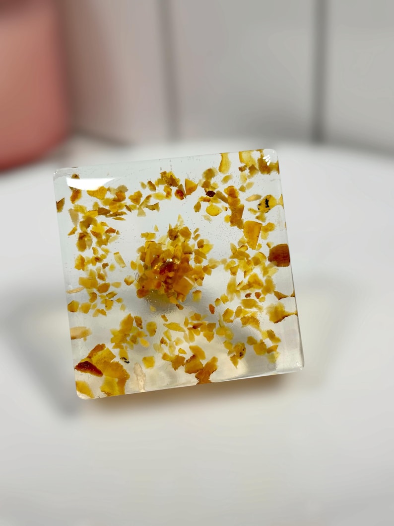 Op de afbeelding: Een vierkant, helder harsblok of presse-papier met ingebedde goudgele, barnsteenachtige fragmenten. De kleine, onregelmatige stukjes zijn verspreid door het transparante materiaal, met een dichtere concentratie in het midden.