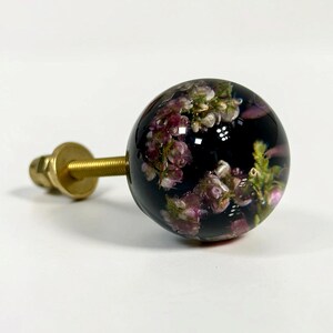 Pode incluir: Um puxador esf&eacute;rico para m&oacute;veis com corpo em resina escura e transl&uacute;cida, incrustado com pequenas flores cor-de-rosa e brancas. Um parafuso e uma anilha em metal dourado sobressaem da parte de tr&aacute;s da esfera para a sua fixa&ccedil;&atilde;o.