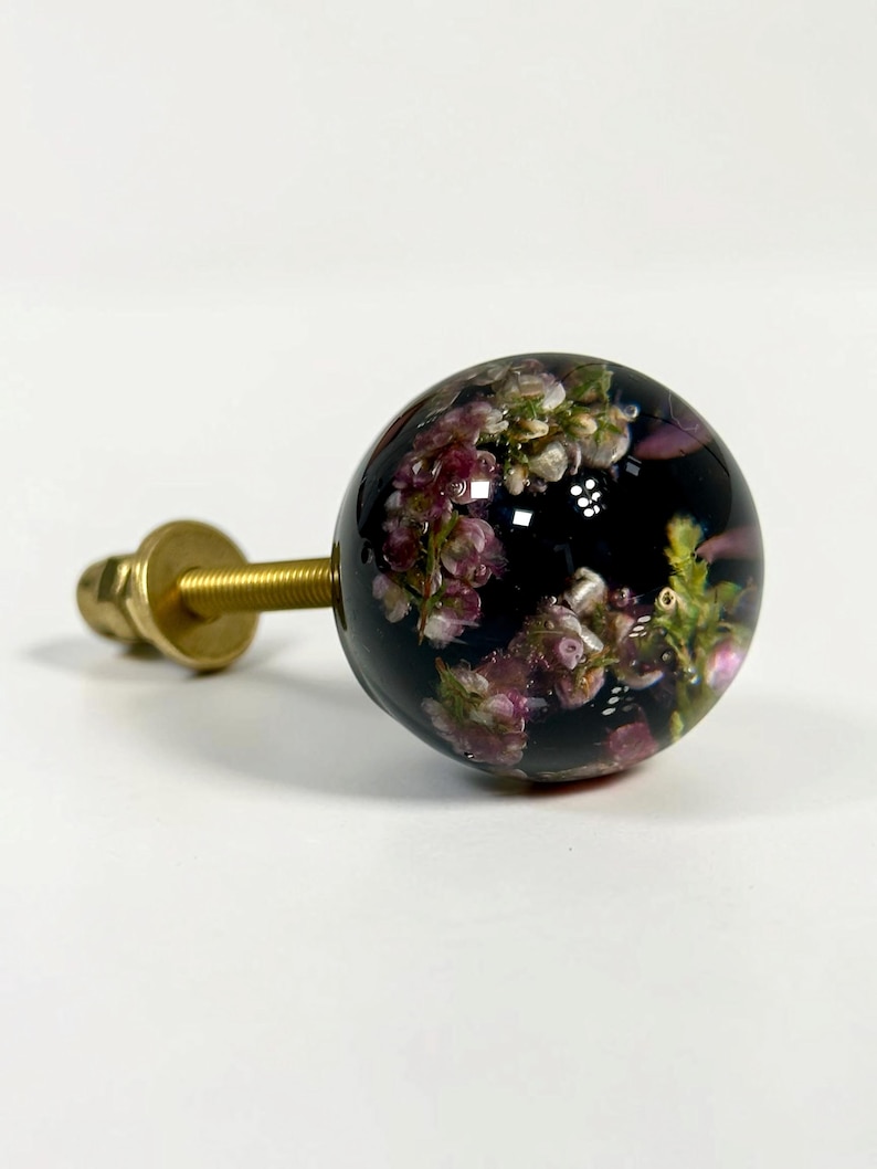 Pode incluir: Um puxador esf&eacute;rico para m&oacute;veis com corpo em resina escura e transl&uacute;cida, incrustado com pequenas flores cor-de-rosa e brancas. Um parafuso e uma anilha em metal dourado sobressaem da parte de tr&aacute;s da esfera para a sua fixa&ccedil;&atilde;o.