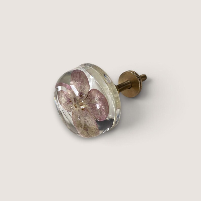 Puede incluir: Un tirador de mueble redondo hecho de resina transparente con una flor de hortensia morada prensada y seca en su interior. El tirador cuenta con una base de tornillo roscado de tono dorado met&aacute;lico para su fijaci&oacute;n, siendo un herraje funcional.