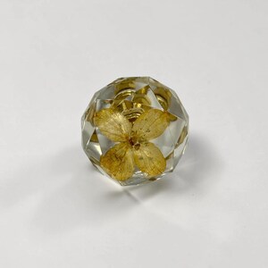 May include: A faceted, clear resin cabinet knob featuring a single, pressed yellow flower encased within the transparent material. The knob has a geometric, multi-sided shape and is attached to a circular, metallic gold-toned base hardware.