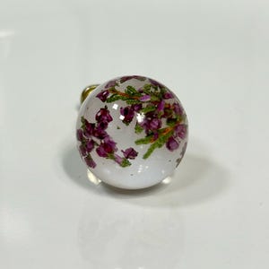 May include: A spherical clear resin charm containing small, dried purple heather flowers and green stems. The object is round and smooth, featuring a metallic attachment point visible at the top, showcasing the botanical elements suspended inside.
