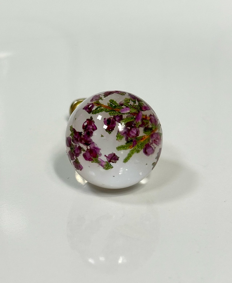 May include: A spherical clear resin charm containing small, dried purple heather flowers and green stems. The object is round and smooth, featuring a metallic attachment point visible at the top, showcasing the botanical elements suspended inside.