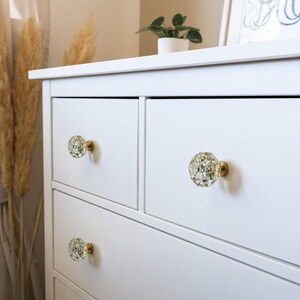 May include: A white dresser featuring three clear, multifaceted, geometric drawer knobs with embedded dried white flowers and gold-toned metal bases. The knobs have a distinct hexagonal shape and are attached to the front of the white drawers.