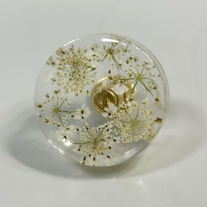 Puede incluir: Un pomo circular para muebles hecho de resina transparente con flores blancas prensadas incrustadas. El pomo tiene una base de metal dorado con un mecanismo de tornillo central visible en el medio, que sirve como punto de fijaci&oacute;n funcional.