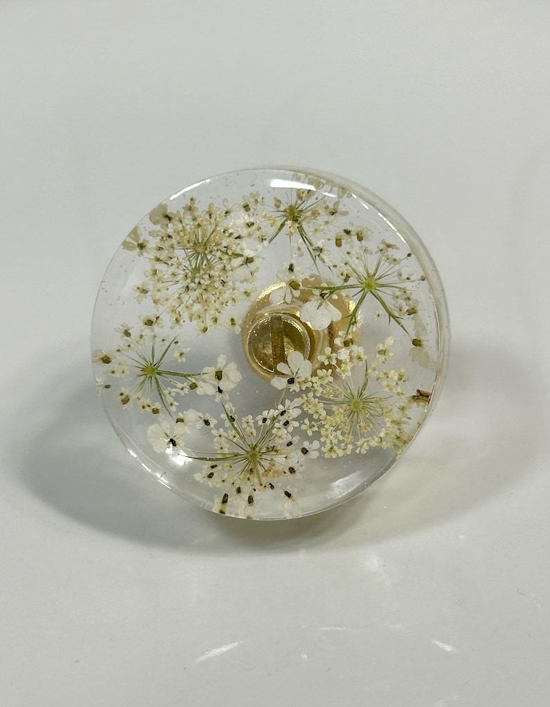 Puede incluir: Un pomo circular para muebles hecho de resina transparente con flores blancas prensadas incrustadas. El pomo tiene una base de metal dorado con un mecanismo de tornillo central visible en el medio, que sirve como punto de fijaci&oacute;n funcional.