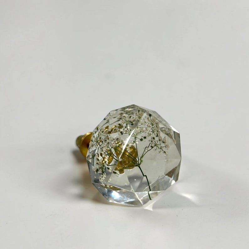 May include: A clear, faceted, multi-sided resin cabinet knob containing a delicate, dried white flower specimen suspended within the material. The knob features a gold-toned metal base attachment extending from the back of the clear structure.