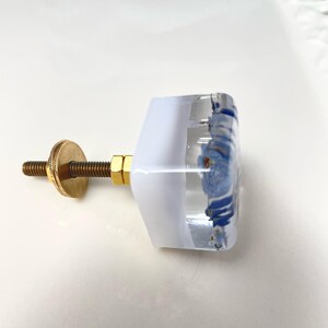 Puede incluir: Un tirador de mueble cuadrado con cuerpo de resina transparente y blanca que contiene motivos florales azules incrustados. El herraje incluye un perno de metal color lat&oacute;n, una tuerca hexagonal y una arandela circular de fijaci&oacute;n.
