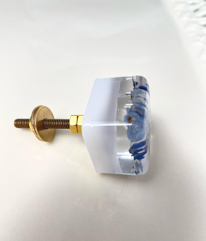 Puede incluir: Un tirador de mueble cuadrado con cuerpo de resina transparente y blanca que contiene motivos florales azules incrustados. El herraje incluye un perno de metal color lat&oacute;n, una tuerca hexagonal y una arandela circular de fijaci&oacute;n.