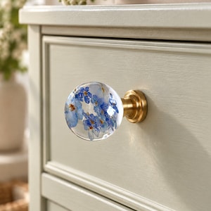 Peut inclure: Un bouton de meuble sphérique composé d'un dôme en résine transparente incrusté de petites fleurs myosotis bleues, fixé sur une base circulaire en laiton brossé. Le bouton est monté sur un tiroir en bois vert sauge clair mouluré.