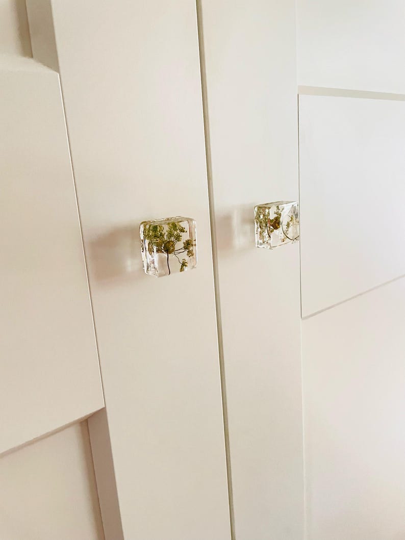 Puede incluir: Dos pomos de armario cuadrados hechos de resina transparente, cada uno con un peque&ntilde;o esp&eacute;cimen bot&aacute;nico verde seco en su interior. Los pomos est&aacute;n fijados a una puerta de armario blanca, lisa y de panel plano, creando un contraste visual.
