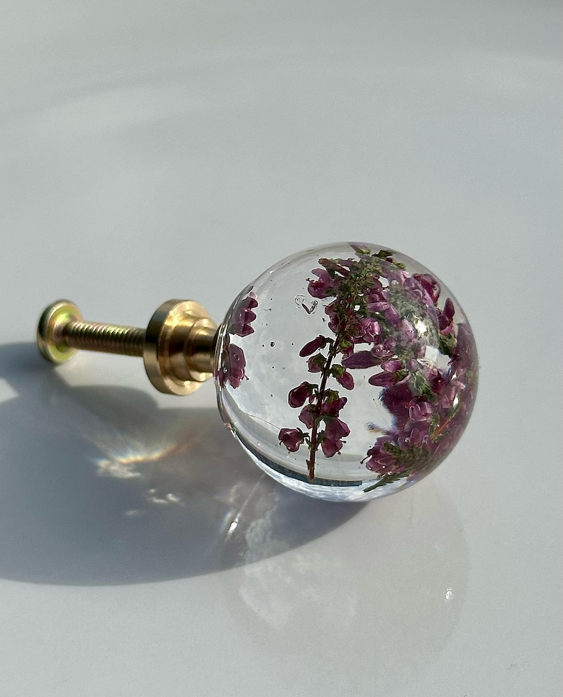 K&ouml;nnte beinhalten: Ein kugelf&ouml;rmiger M&ouml;belknopf aus klarem Kunstharz mit eingebetteten violetten Heidekrautbl&uuml;ten. Der Knopf ist an einer goldfarbenen Metallschraube mit Sockel befestigt und dient als funktionales Beschlagteil f&uuml;r M&ouml;bel, Schubladen oder T&uuml;ren.