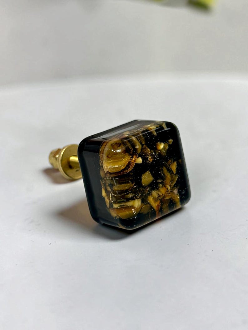K&ouml;nnte beinhalten: Ein quadratischer M&ouml;belknopf mit gl&auml;nzender schwarzer Harzoberfl&auml;che, in die schimmernde goldene und braune Partikel eingebettet sind. Der Knopf ist an einem runden, messingfarbenen Metallsockel mit kurzem Gewindebolzen befestigt.
