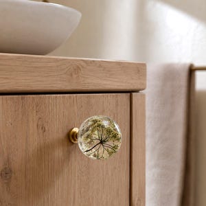May include: A spherical cabinet knob made of clear resin featuring an embedded dried white flower. The knob is attached to a light wood cabinet door with a circular brass-toned metal base, providing a secure and functional grip.