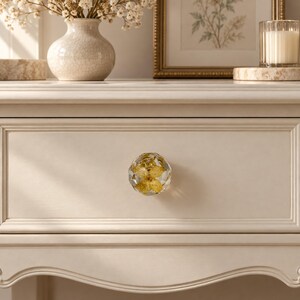 May include: A round, faceted clear resin drawer knob featuring an embedded yellow dried flower. The knob is attached to a cream-colored wooden drawer front with a smooth, matte finish and decorative molding along the edges of the drawer.