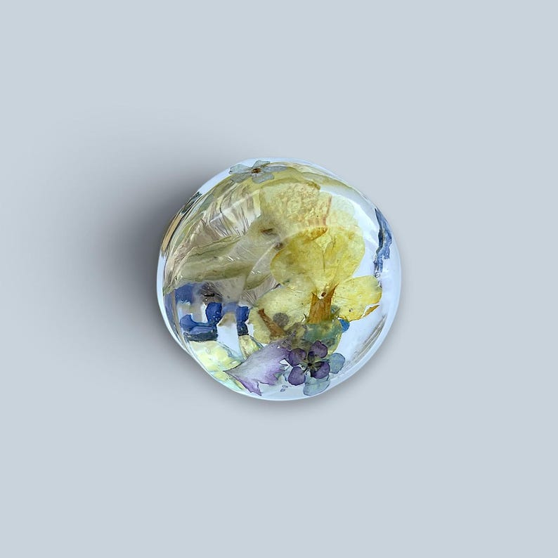 May include: A circular, clear resin cabochon containing pressed botanical elements. The embedded flowers include yellow petals, small blue blossoms, and light purple accents, all suspended within the transparent, smooth, and rounded material.