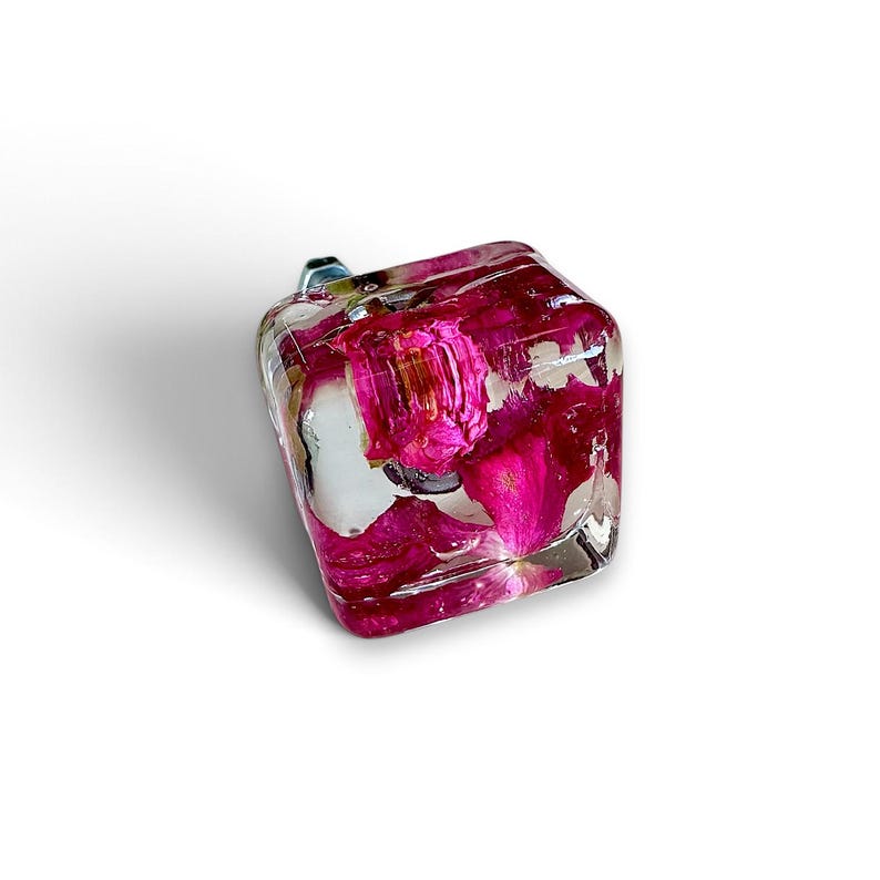 May include: A square cabinet knob made of clear resin with embedded vibrant pink flower petals. The knob features a smooth, glossy finish and a metallic mounting screw visible at the back, providing a functional hardware piece for furniture.