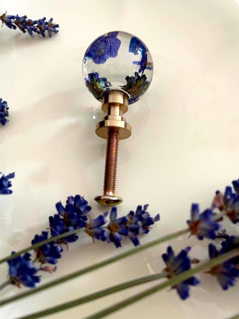 May include: A spherical cabinet knob made of clear resin containing embedded blue flowers. The knob features a metallic gold-toned base and a long threaded metal bolt extending from the bottom, surrounded by sprigs of dried purple lavender.