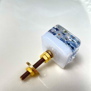Puede incluir: Un tirador de mueble cuadrado con una parte superior de resina transparente que contiene peque&ntilde;as flores azules y una base blanca s&oacute;lida. Incluye un perno met&aacute;lico dorado, dos tuercas doradas a juego y una arandela plana para montaje.