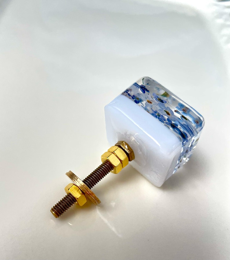 Puede incluir: Un tirador de mueble cuadrado con una parte superior de resina transparente que contiene peque&ntilde;as flores azules y una base blanca s&oacute;lida. Incluye un perno met&aacute;lico dorado, dos tuercas doradas a juego y una arandela plana para montaje.