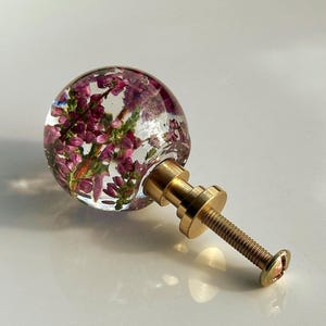 K&ouml;nnte beinhalten: Ein kugelf&ouml;rmiger M&ouml;belknopf aus klarem Harz, in das kleine violette Heidekrautbl&uuml;ten eingebettet sind. Der Knopf ist an einem goldfarbenen Metallsockel mit einer Gewindeschraube befestigt und dient als funktionales Beschlagteil.