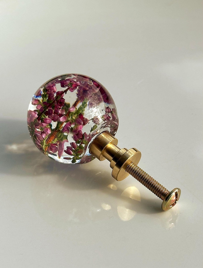 K&ouml;nnte beinhalten: Ein kugelf&ouml;rmiger M&ouml;belknopf aus klarem Harz, in das kleine violette Heidekrautbl&uuml;ten eingebettet sind. Der Knopf ist an einem goldfarbenen Metallsockel mit einer Gewindeschraube befestigt und dient als funktionales Beschlagteil.