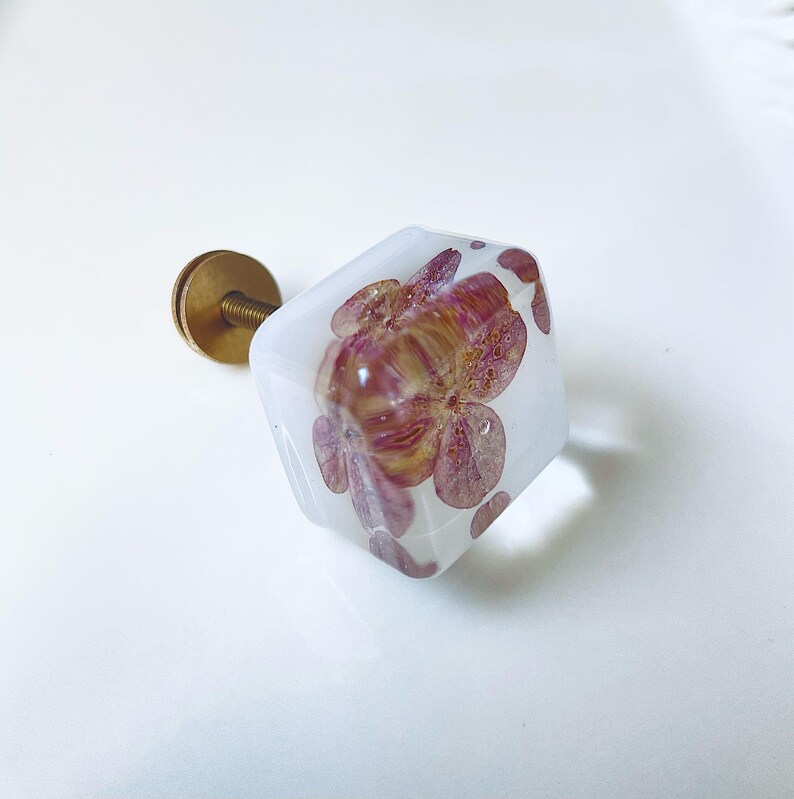 May include: A square cabinet knob made of clear resin with embedded dried pink and purple hydrangea petals. The knob features a brass-colored metal screw and base attachment extending from the back, providing a functional mounting point.