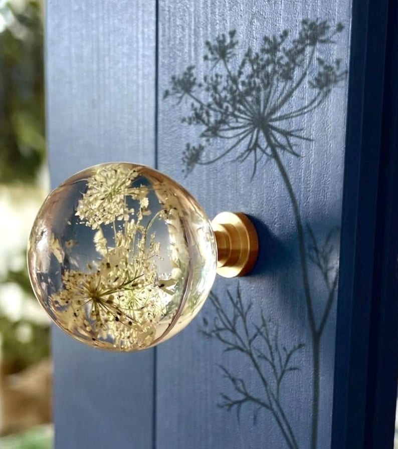 May include: A spherical clear resin cabinet knob containing embedded white dried flowers. The knob features a polished gold-tone metal base and is mounted onto a vertical dark blue wooden panel, casting a distinct shadow of the floral design.