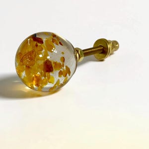 Puede incluir: Un pomo esf&eacute;rico para muebles con un cuerpo de resina transparente incrustado con peque&ntilde;os fragmentos irregulares de color &aacute;mbar. El pomo est&aacute; unido a un perno de metal dorado con una arandela y una tuerca a juego.