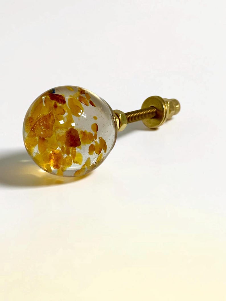 Puede incluir: Un pomo esf&eacute;rico para muebles con un cuerpo de resina transparente incrustado con peque&ntilde;os fragmentos irregulares de color &aacute;mbar. El pomo est&aacute; unido a un perno de metal dorado con una arandela y una tuerca a juego.