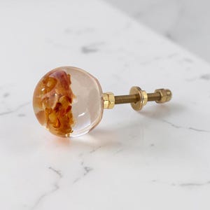 Puede incluir: Un pomo esf&eacute;rico para muebles hecho de resina transparente con fragmentos org&aacute;nicos de color &aacute;mbar incrustados. El pomo cuenta con un perno de metal dorado con una tuerca y arandela a juego para su fijaci&oacute;n.