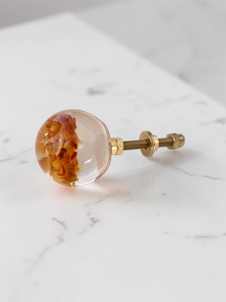 Puede incluir: Un pomo esf&eacute;rico para muebles hecho de resina transparente con fragmentos org&aacute;nicos de color &aacute;mbar incrustados. El pomo cuenta con un perno de metal dorado con una tuerca y arandela a juego para su fijaci&oacute;n.