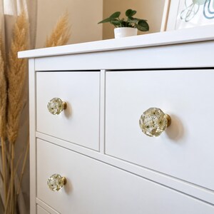 May include: A white dresser featuring three round, faceted drawer knobs made of clear resin. Each knob contains embedded dried white flowers and is attached to the drawer front with a circular brass-colored metal base mount.