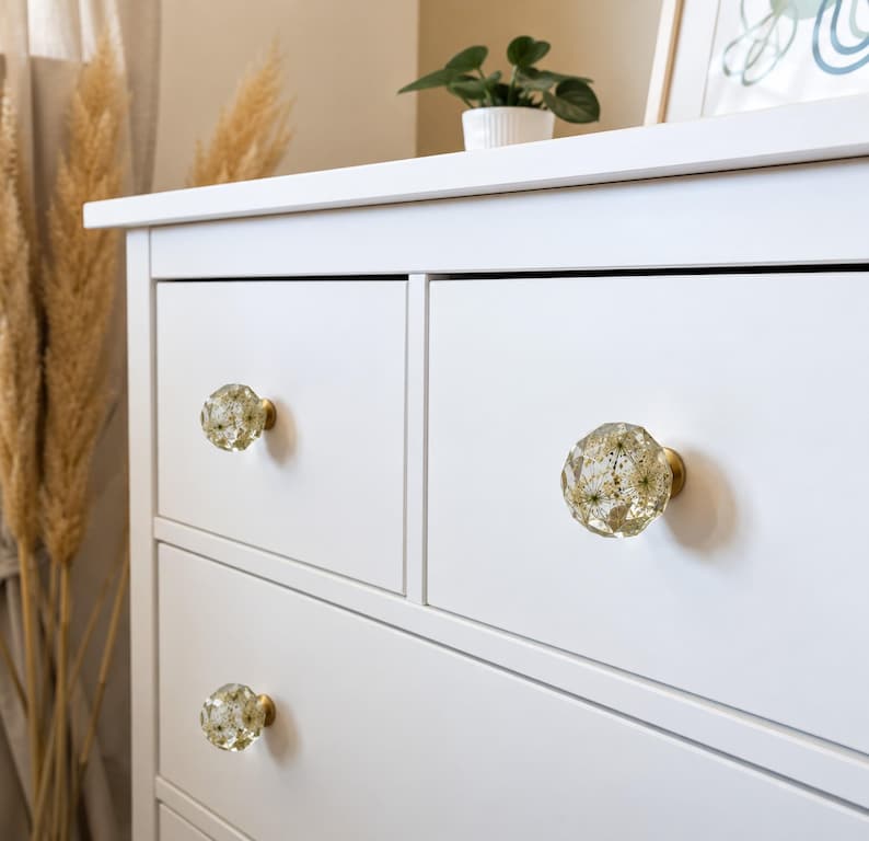 May include: A white dresser featuring three round, faceted drawer knobs made of clear resin. Each knob contains embedded dried white flowers and is attached to the drawer front with a circular brass-colored metal base mount.