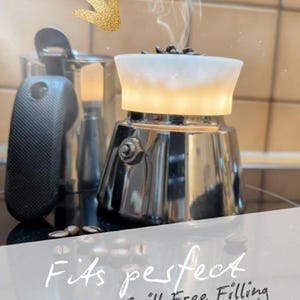 Könnte beinhalten: Ein metallischer Espressokocher mit einem weißen Trichteraufsatz, in dem sich Kaffeebohnen befinden und aus dem Dampf aufsteigt. Der Text auf dem Bild lautet: Fits perfect. Effortless, Spill-Free Filling. Ein goldener Pfeil zeigt auf den Trichter.