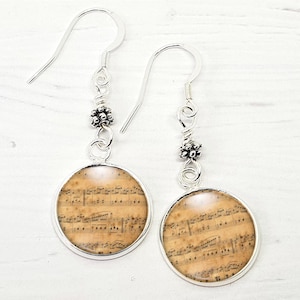 May include: Silver drop earrings with circular pendants displaying vintage sheet music. The earrings have a simple hook design and decorative bead accents. The musical notes are printed on a cream-coloured background.