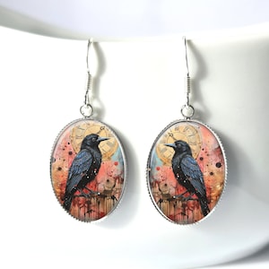 May include: A pair of silver-toned dangle earrings. Each earring features an oval pendant with a black crow illustration perched on a branch, set against a clock face and abstract background. The earrings are suspended from a curved hook.